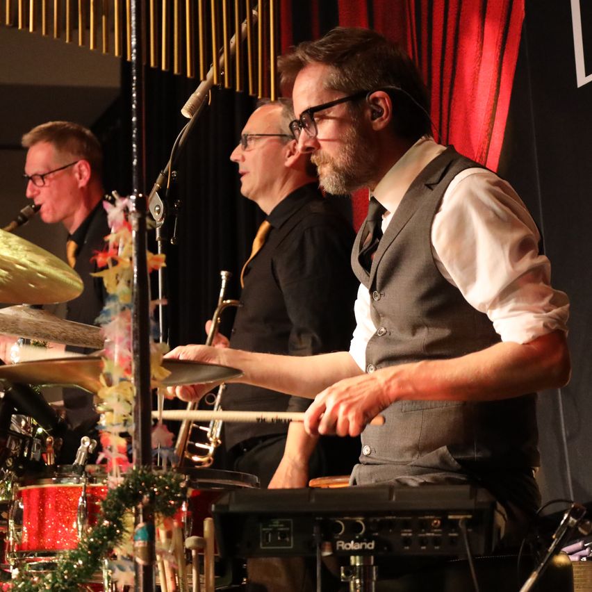 Drei Musiker treten auf der Bühne auf. Der Mann im Vordergrund spielt die Trommeln. Der Mann hinter ihm spielt die Trompete. Der Mann auf der linken Seite spielt das Saxophon.