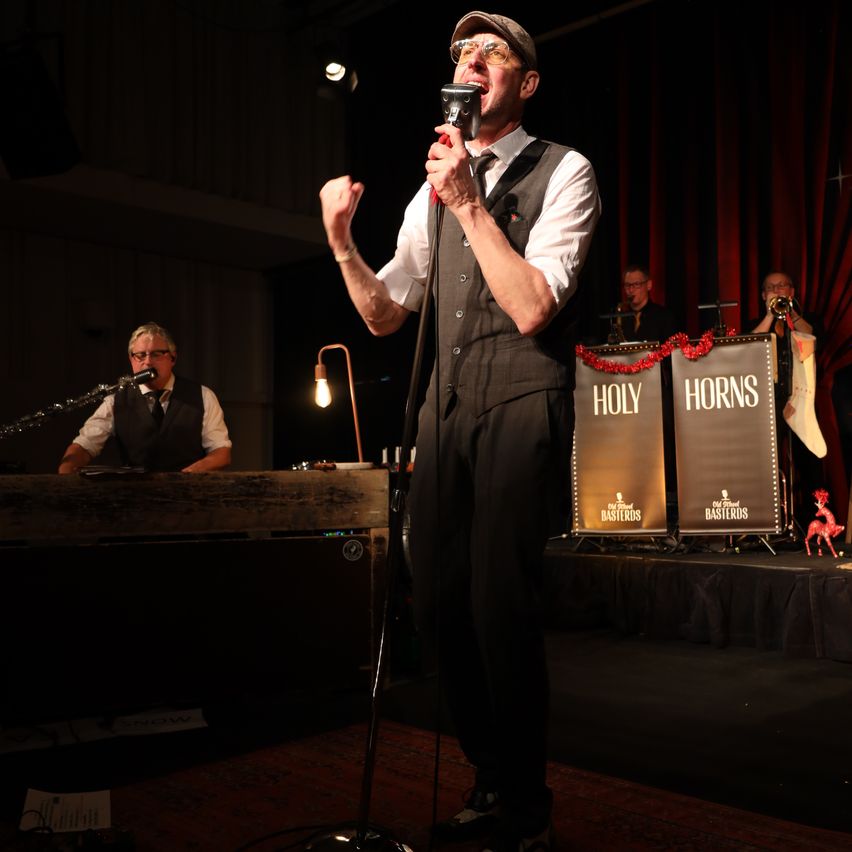 Ein Mann singt auf der Bühne mit einem Mikrofon, eine Band dahinter und ein Hintergrund mit dem Text Holy Horns.