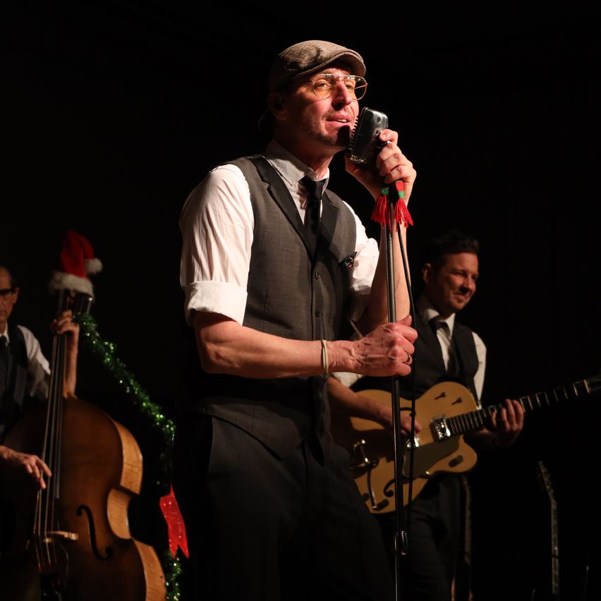 Ein Mann singt in ein Mikrofon auf der Bühne mit einer Band. Er trägt eine Kappe und eine Brille. Hinter ihm spielt ein Cellist, während ein Gitarrist spielt. Die Bühne ist dunkel.