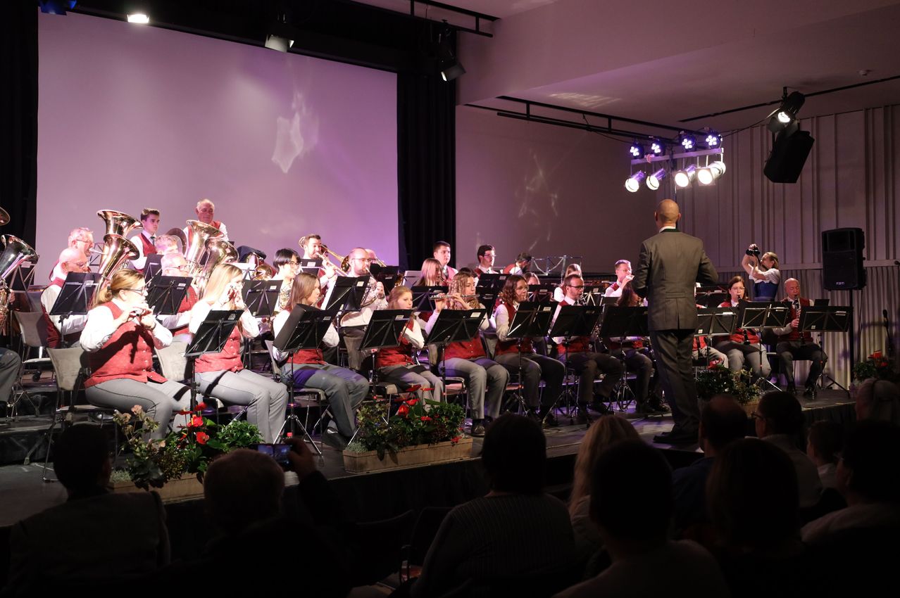 Ein Orchester spielt auf der Bühne, mit Musikern, die sitzen und verschiedene Instrumente spielen. Das Publikum sitzt davor und beobachtet die Aufführung. Die Bühne ist gut beleuchtet mit Scheinwerfern.