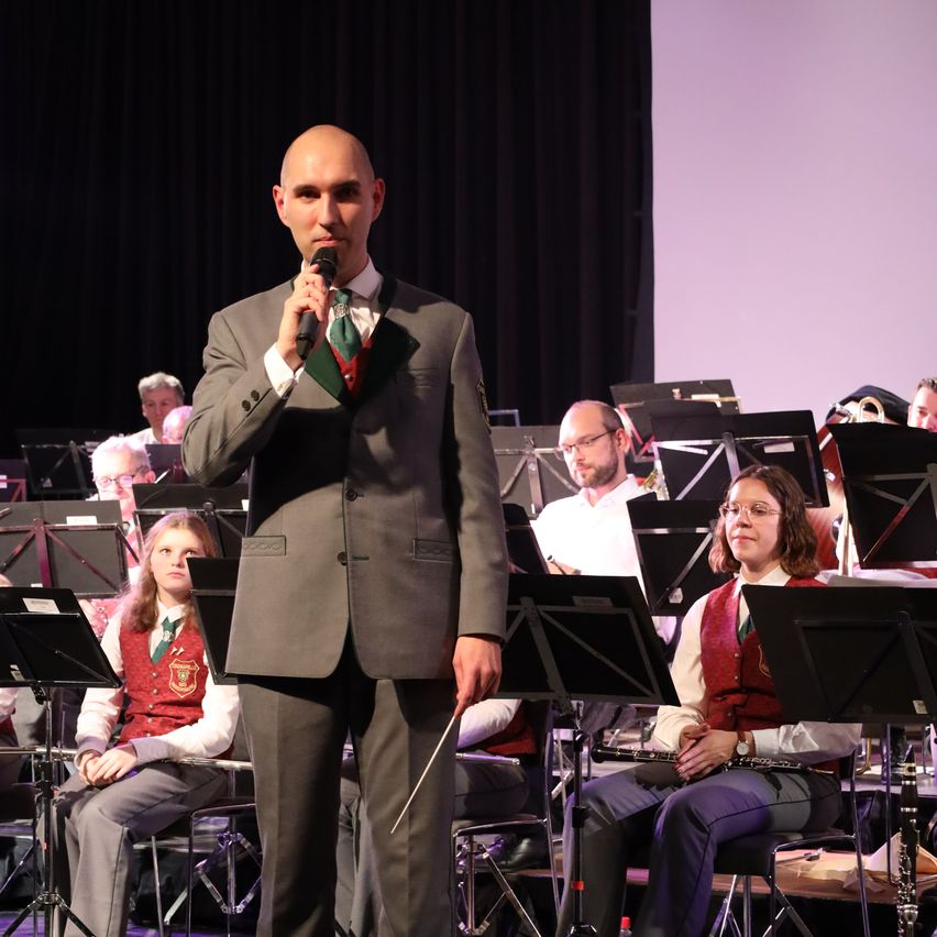 Ein Mann in Anzug und Krawatte singt und hält ein Mikrofon, während er vor einem sitzenden Orchester steht.