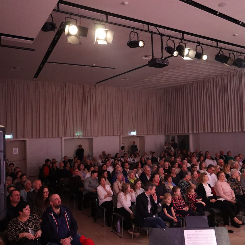 Ein großer Saal mit einem Publikum, das in Reihen sitzt und zur Bühne blickt. Scheinwerfer hängen von der Decke, und eine Person steht in der Nähe der Bühne. Das Publikum ist vielfältig, mit verschiedenen Kleidungsstilen.