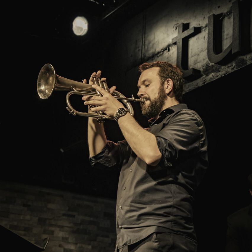 Ein Mann spielt Trompete auf einer Bühne mit Ziegelsteinwänden im Hintergrund und Scheinwerfern, die den Bereich beleuchten.