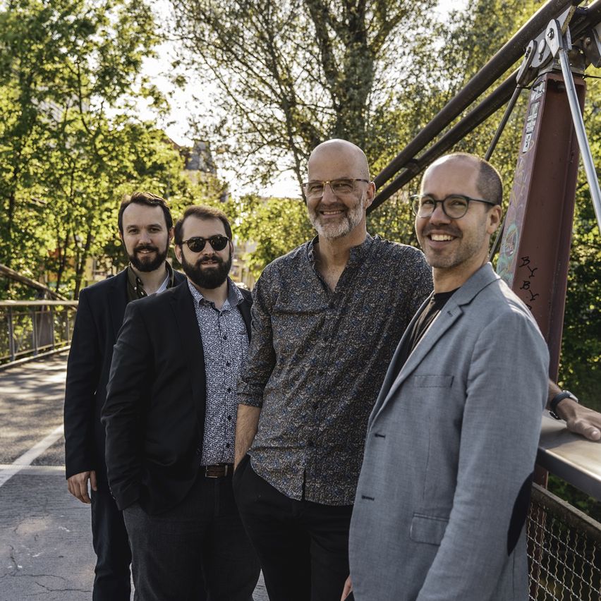Vier Männer stehen auf einer Brücke und lächeln für ein Foto. Hinter ihnen befinden sich Bäume und ein Metallgeländer. Der Mann auf der linken Seite trägt eine Sonnenbrille. Der Mann in der Mitte hat einen Bart und eine Brille. Der Mann auf der rechten Seite trägt einen grauen Anzug.