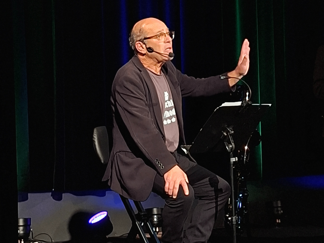 Ein älterer Mann mit Brille sitzt auf einem Stuhl und hält eine Rede mit einem Mikrofon auf einem Ständer. Er gestikuliert mit einer Hand, während die andere auf seinem Schoß ruht.