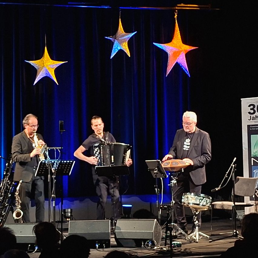 Drei Musiker spielen auf der Bühne mit Saxophon, Akkordeon und Schlagzeug. Drei Sterndekorationen hängen über ihnen. Ein Publikum sitzt vor der Bühne.