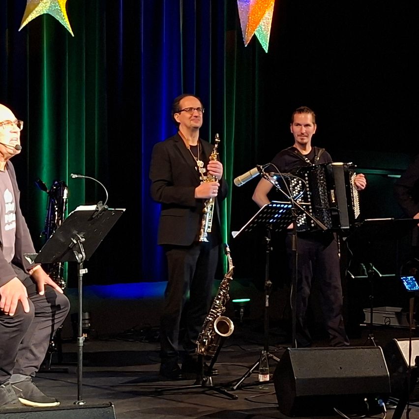 Drei Musiker stehen auf der Bühne und spielen. Einer sitzt und spielt Saxophon, während die anderen beiden stehen, einer mit einem Saxophon und der andere mit einem Akkordeon. Mikrofone und Notenständer stehen vor ihnen.