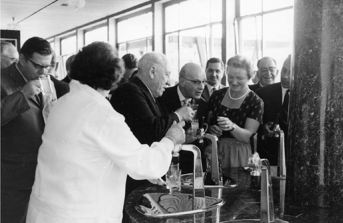 Ein altes Foto zeigt eine Gruppe von Menschen in einer Bar, ein Mann schenkt ein Getränk ein, andere beobachten.
