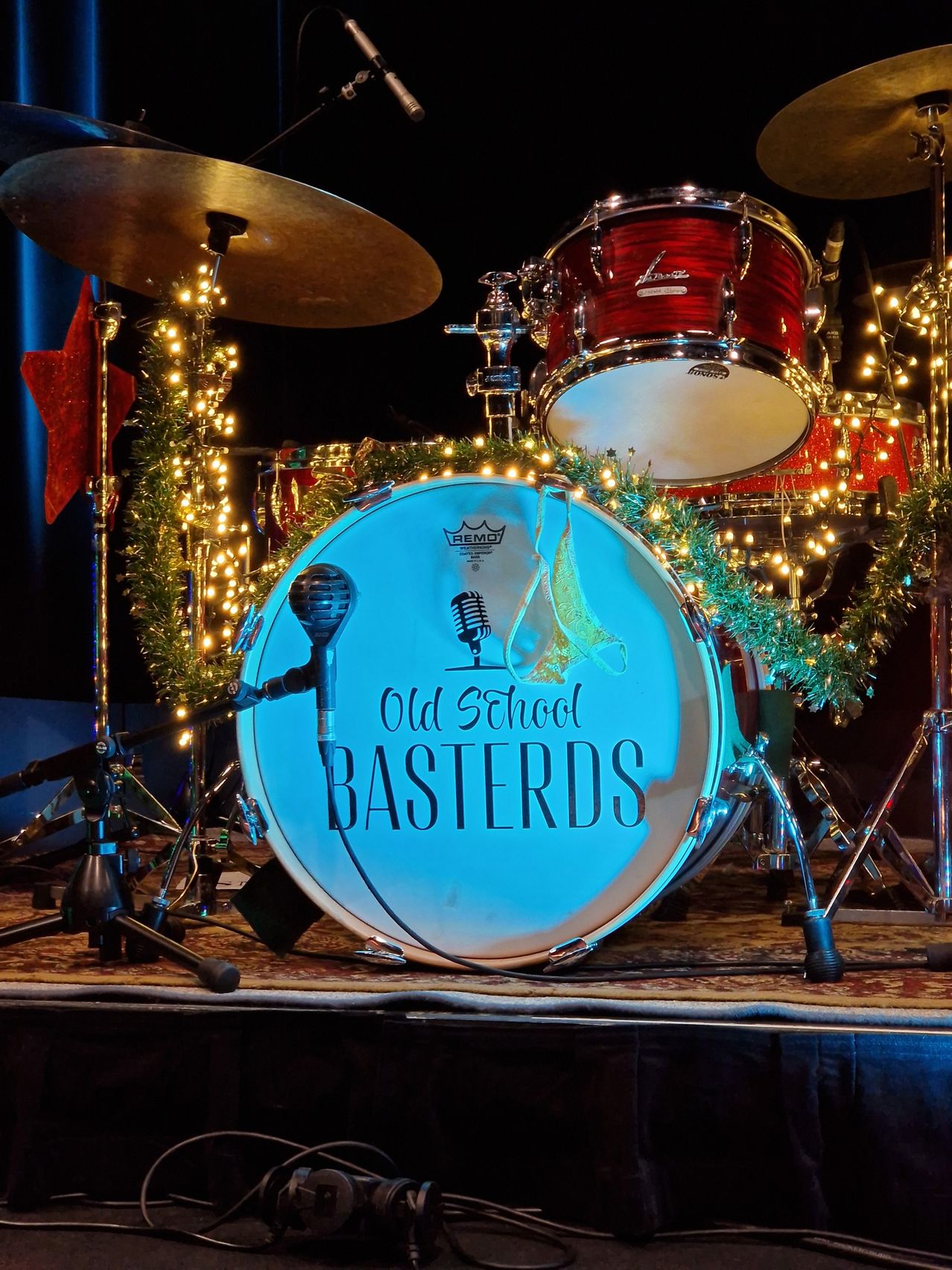 A drum set on stage, decorated with Christmas lights, featuring the word 'Old School Bastards' on the bass drum.