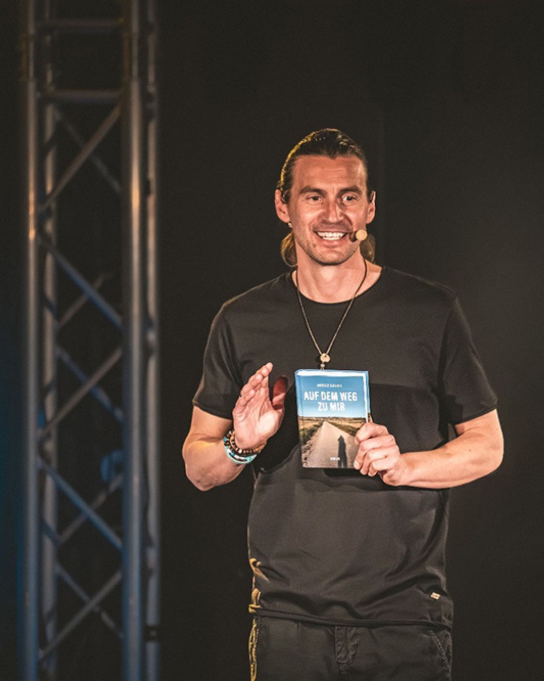 A man with a microphone and a necklace stands on a stage, smiling, and holding a book titled 'AUF DEM WEG ZU MIR'. He appears to be speaking to an audience.