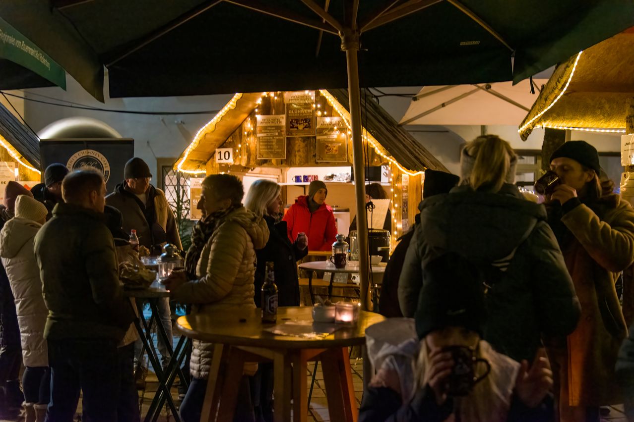 Eine Gruppe von Menschen versammelt sich unter einem Regenschirm bei einer festlichen Veranstaltung im Freien. Die Szene ist von Lichterketten beleuchtet, und eine Frau in einer roten Jacke steht in der Nähe eines Tisches.