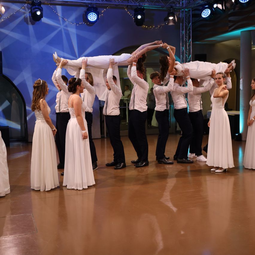 A group of people in formal attire are performing a lift with two women in white dresses on a wooden floor. They are all smiling.