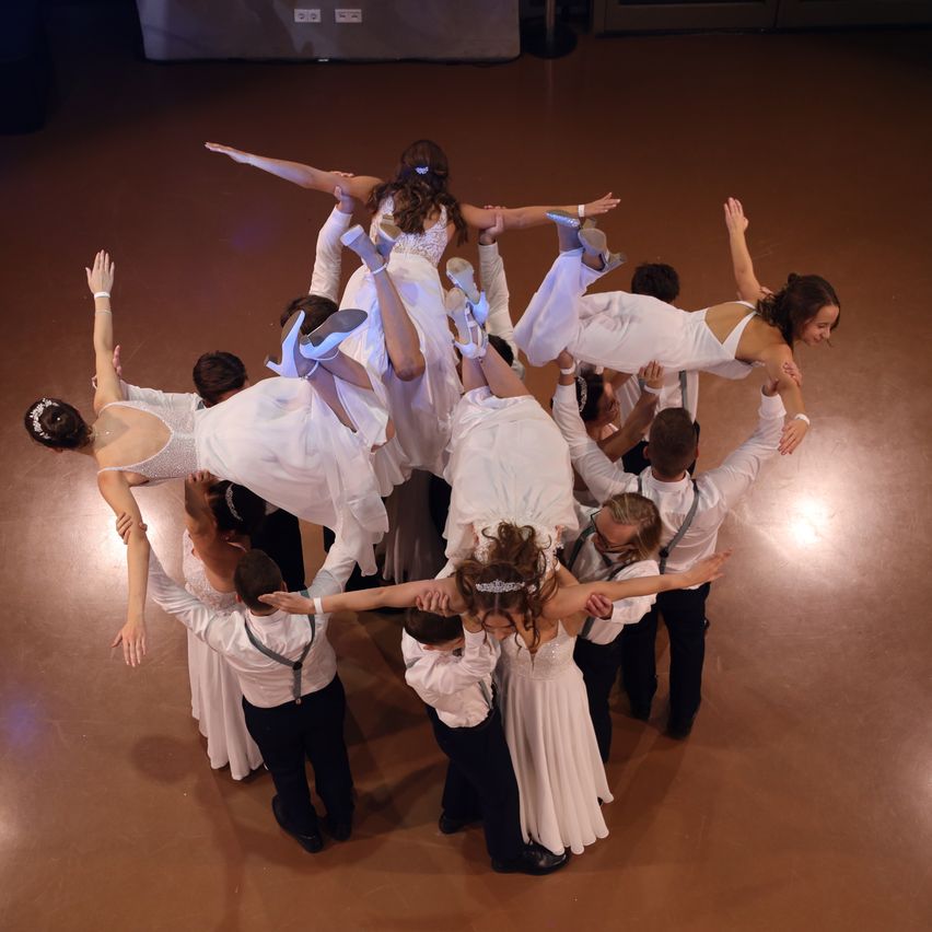 A group of dancers, including several women in white dresses, are performing a coordinated lift on a dance floor. The dancers are wearing white dresses and white shoes, with some wearing bracelets.