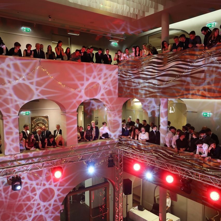 A large group of people are gathered indoors. They are standing on multiple levels, with some on balconies. The room is decorated with pink lighting and geometric patterns. Several individuals are looking down towards the main floor.