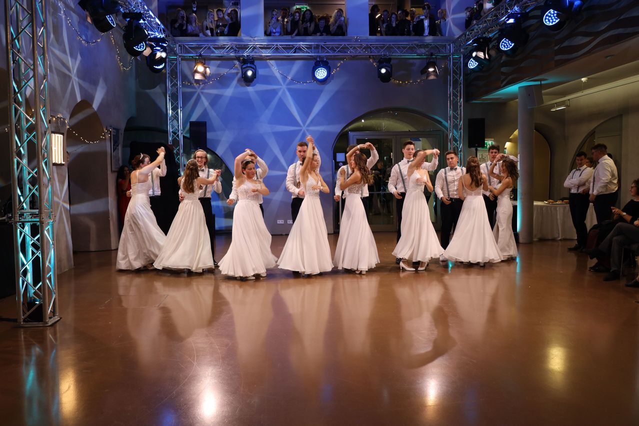 A group of women in white dresses dancing on a stage with blue lights. Spectators are watching from a balcony above.