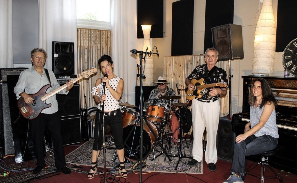 Eine Band tritt in einem Raum auf. Eine Frau singt in ein Mikrofon, während ein Mann Gitarre spielt. Ein Mann spielt Schlagzeug und ein anderer Gitarre. Eine Frau sitzt auf einem Stuhl vor einem Klavier.