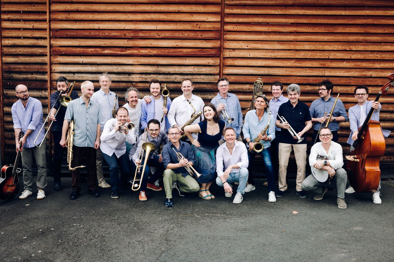 A group of musicians, both male and female, are posing for a picture in front of a wooden wall. They are all holding their musical instruments.