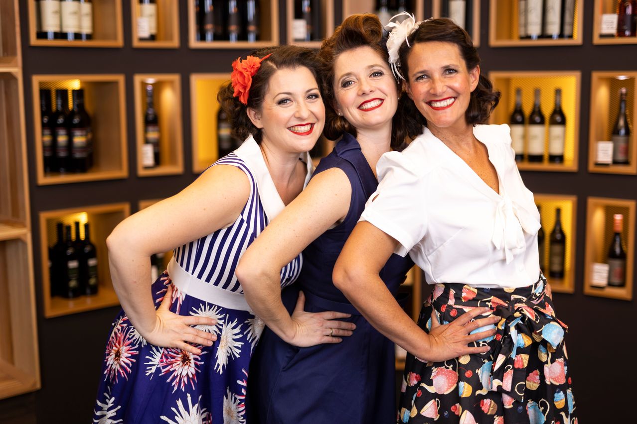 Three women dressed in retro-style dresses are standing together and smiling for a photo in front of a wall with shelves of wine bottles.