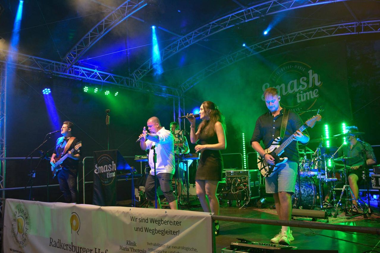 A rock band performs on stage under blue and green lights. A woman and two men are singing, while another man plays the guitar. The stage is adorned with various musical instruments.