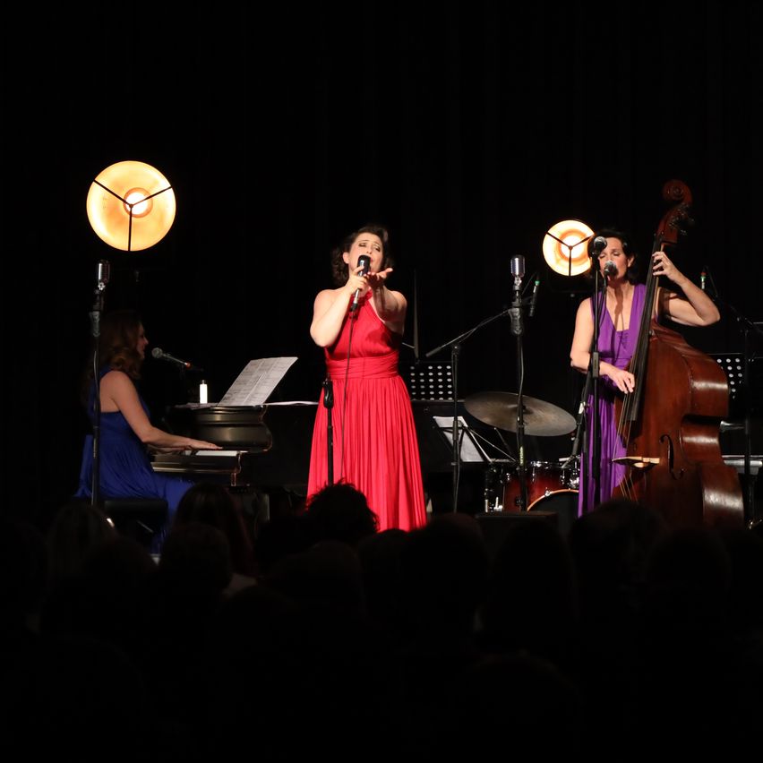 Eine Aufführung auf der Bühne mit drei Frauen. Die Frau in Rot singt in ein Mikrofon, während die anderen zwei Instrumente spielen. Eine spielt Cello und die andere Klavier. Es gibt Mikrofone auf Ständern und eine Scheinwerfer auf der Bühne.