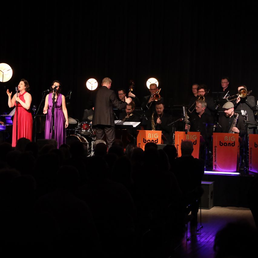 Eine Jazzband tritt mit zwei Sängerinnen auf der Bühne auf. Die Band ist mit orangefarbenen Bannern, die 'BIG Band' lesen, geschmückt. Das Publikum sitzt vor der Bühne, die schwach beleuchtet ist.