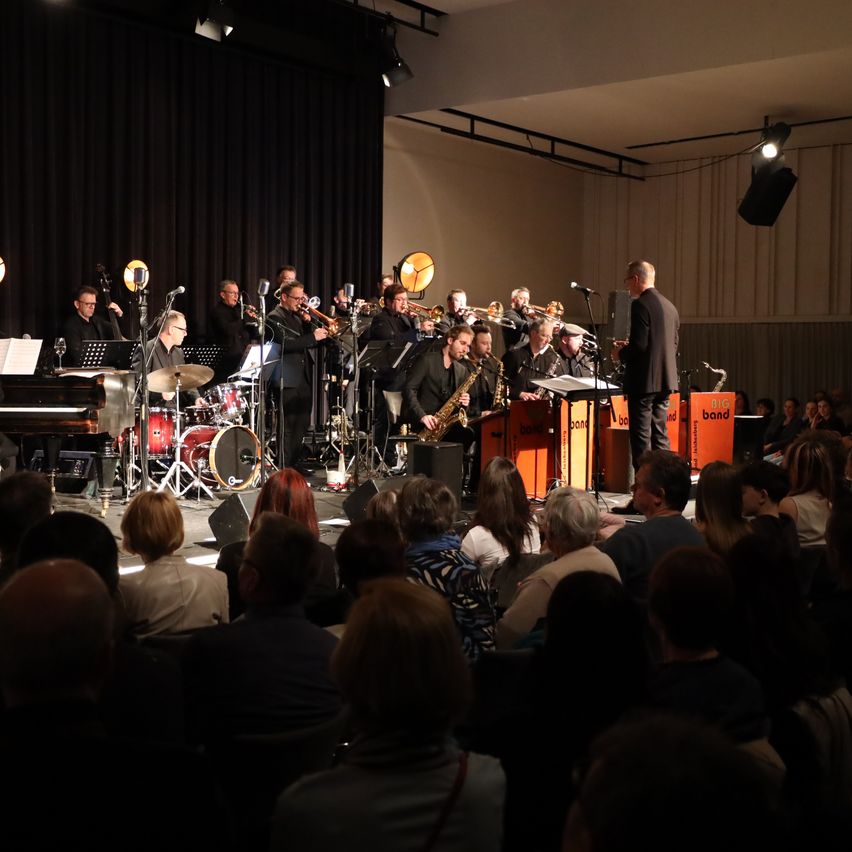 Eine Jazzband spielt auf der Bühne mit verschiedenen Instrumenten, darunter Trompeten, Saxophone und Schlagzeug. Das Publikum sitzt vorne und hört aufmerksam zu. Ein Dirigent steht vorne auf der Bühne und leitet die Musiker.