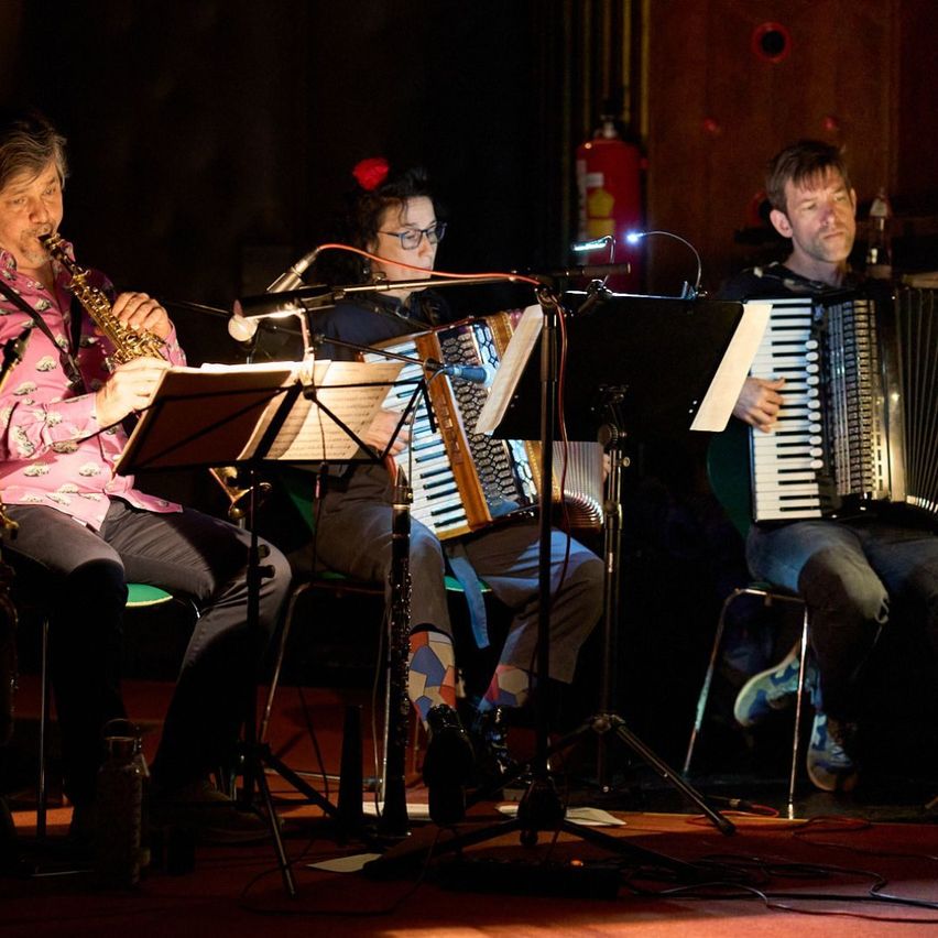 Drei Musiker spielen auf der Bühne mit Saxophon, Akkordeon und Klarinette. Sie sitzen, konzentrieren sich und werden von Bühnenlichtern beleuchtet.