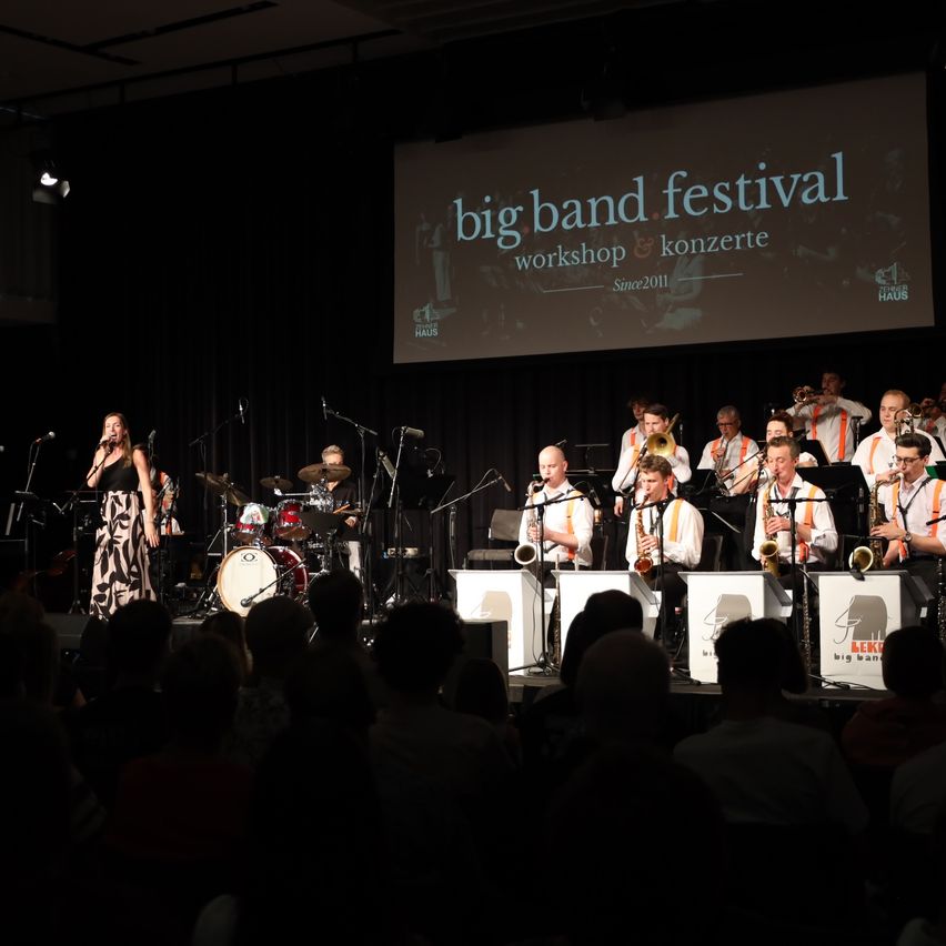 Ein Jazz-Band spielt auf der Bühne, im Hintergrund steht eine große Leinwand mit der Aufschrift 'Big Band Festival'. Eine Sängerin steht vor der Band und singt in ein Mikrofon.