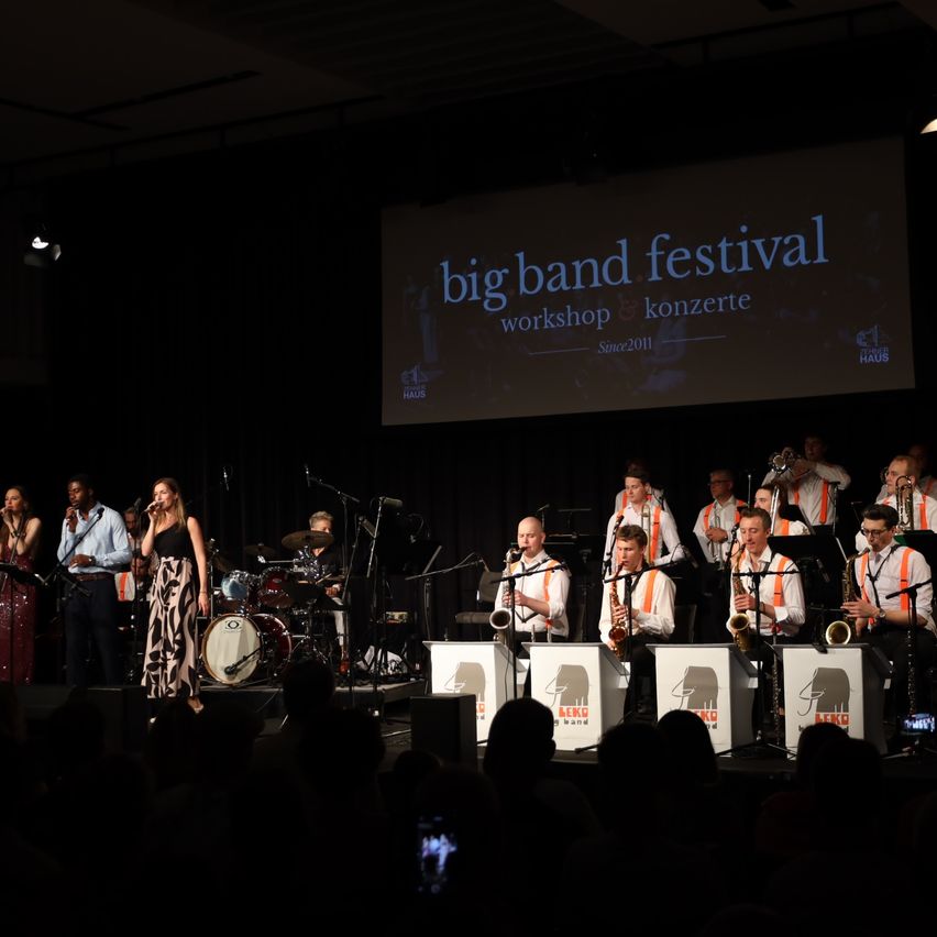 Eine Jazzband tritt auf der Bühne beim Big Band Festival auf, mit einer großen Leinwand im Hintergrund, die den Namen und Details des Festivals anzeigt.