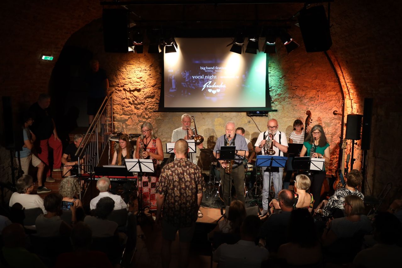 Eine Jazzband spielt auf der Bühne, mit einer Projektionsleinwand hinter ihnen, auf der 'Big Band Festival' und 'Vocal Night' steht. Das Publikum sitzt vor ihnen.