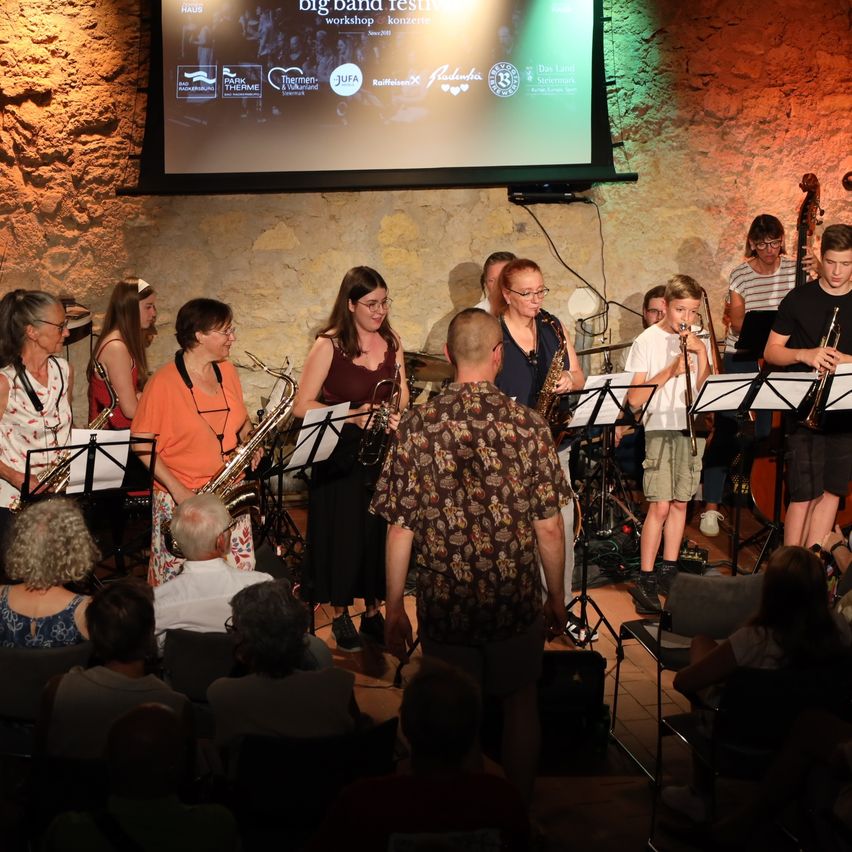 Eine Gruppe Musiker, darunter Kinder, spielen auf der Bühne mit Saxophonen und Trompeten vor einem Publikum. Eine Projektionsleinwand hinter ihnen zeigt den Text 'Big Band Festival'.