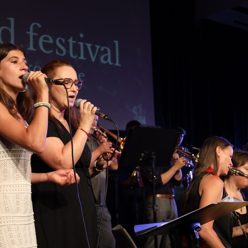 Eine Gruppe von Frauen tritt auf der Bühne bei einem Festival auf, singt in Mikrofone, während eine Band im Hintergrund spielt.