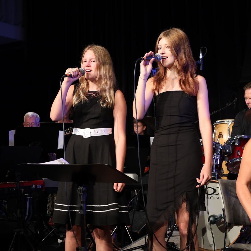 Zwei junge Frauen singen auf der Bühne in Mikrofone. Sie tragen schwarze Kleider. Hinter ihnen spielt eine Band, darunter Schlagzeug und Klavier.