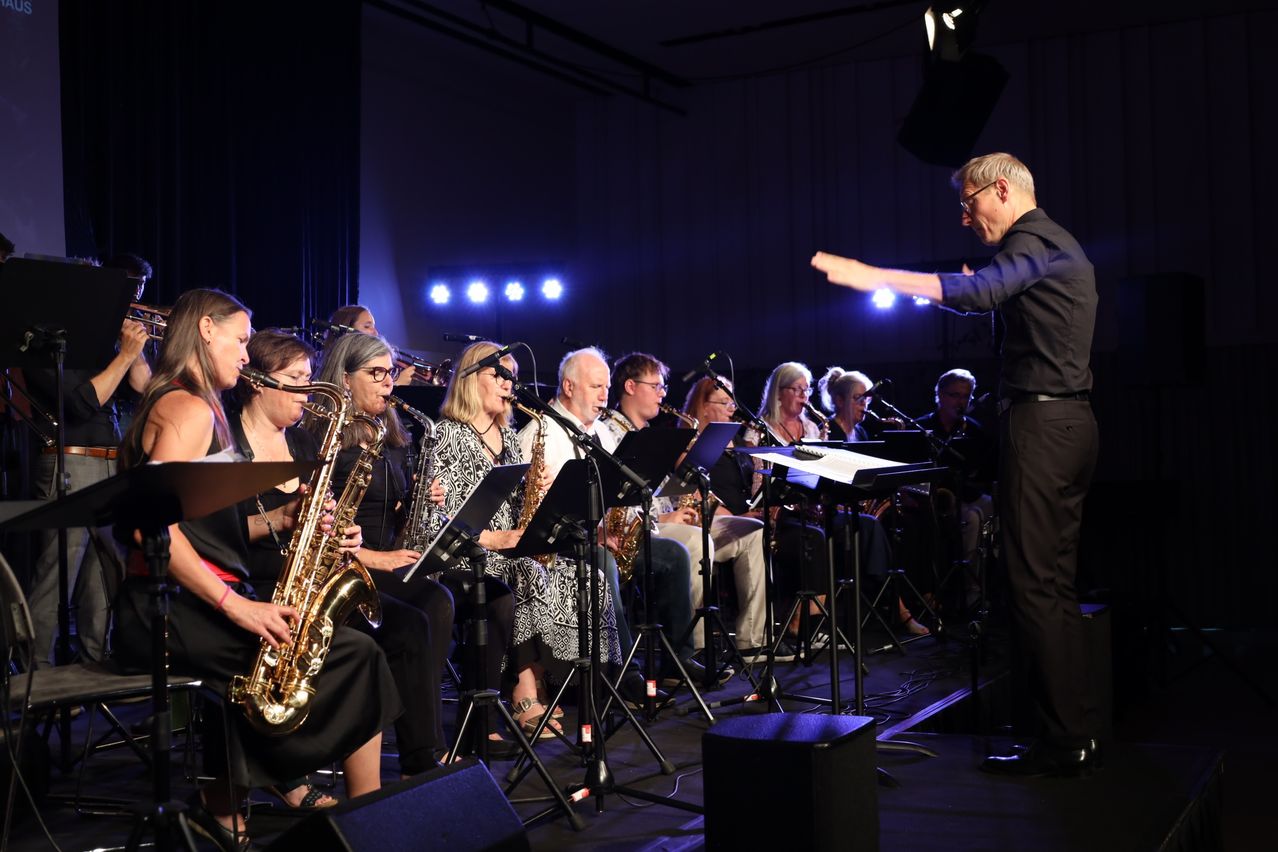 Eine Gruppe von Musikern, einschließlich Saxophonisten, tritt mit einem Dirigenten auf der Bühne auf. Die Bühne ist mit blauen Lichtern gut beleuchtet.