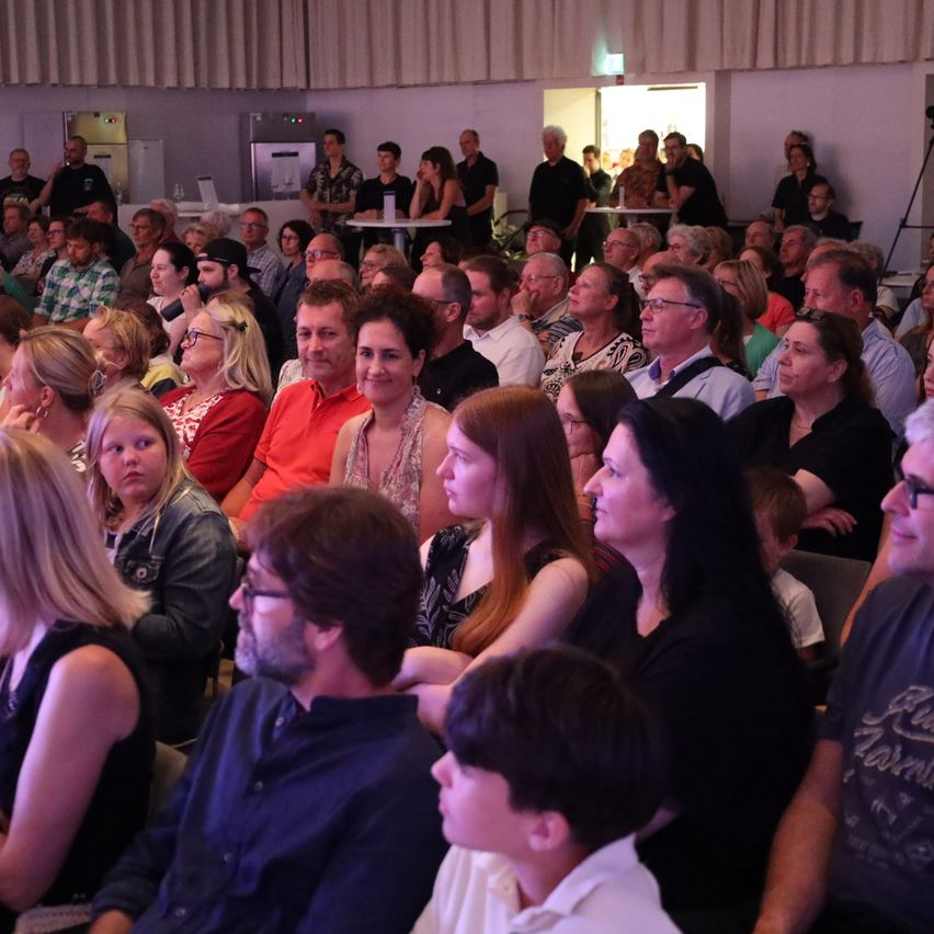 Eine große Gruppe von Menschen sitzt in einem Raum, möglicherweise bei einer Konferenz oder Versammlung. Einige schauen nach vorne, während andere in Gespräche vertieft sind. Der Raum hat eine weiße Wand und einen Vorhang im Hintergrund.