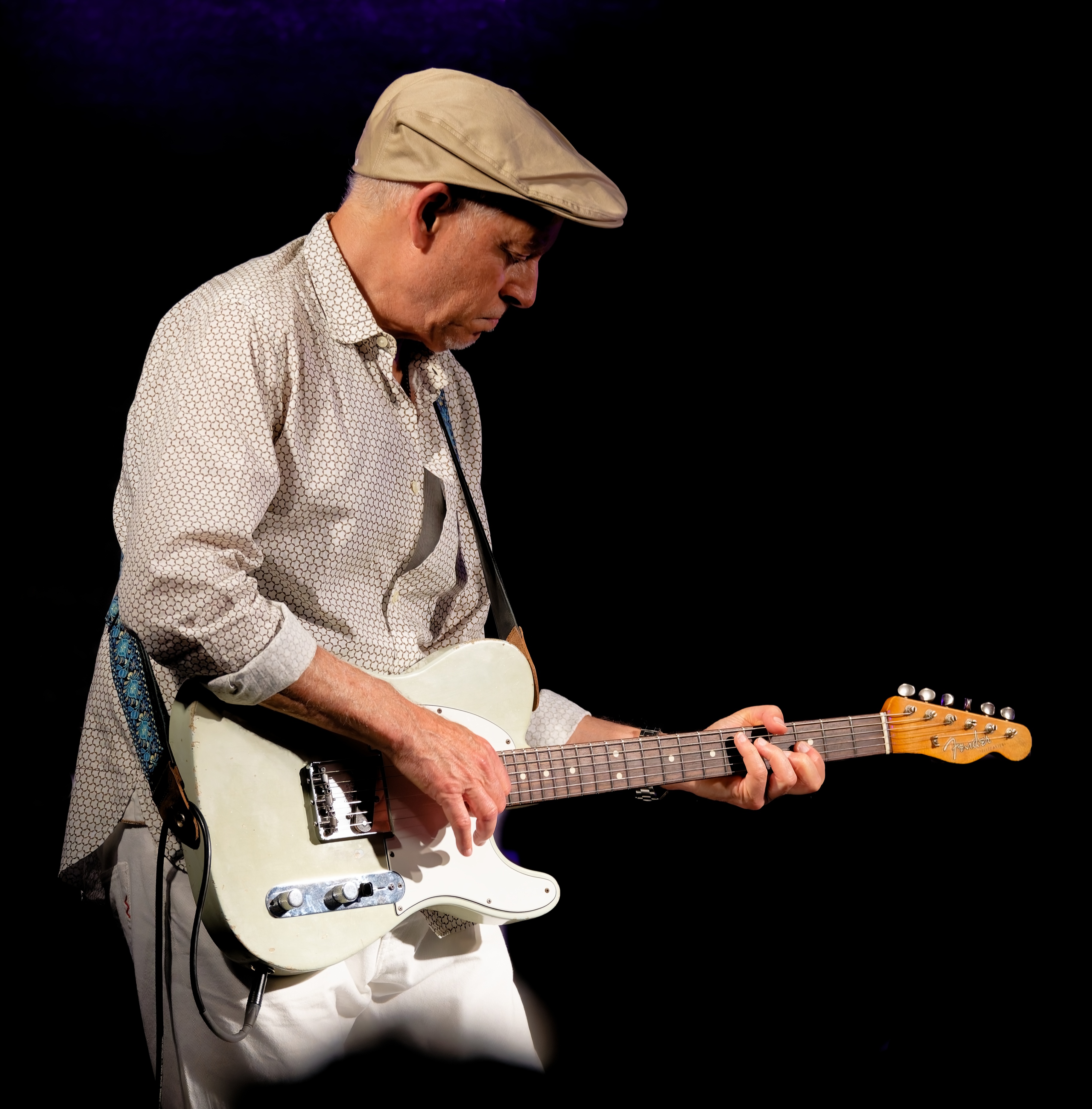 Gitarrist mit Schiebermütze spielt auf weißer E-Gitarre auf dunklem Bühnenhintergrund.