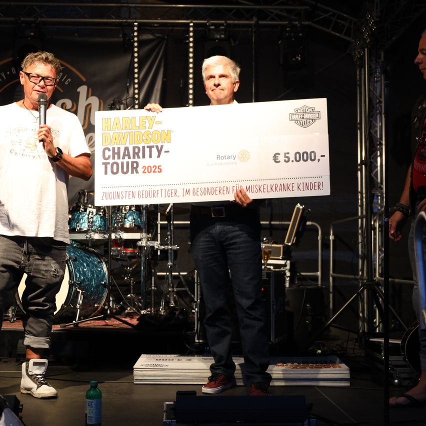 Zwei Männer auf der Bühne halten einen großen Scheck für die Harley-Davidson Charity Tour 2025, einer spricht in ein Mikrofon und der andere hält den Scheck.