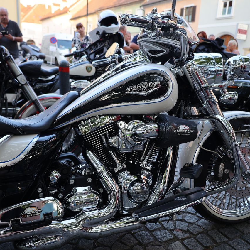 Ein glänzendes schwarzes und silbernes Harley-Davidson Motorrad, geparkt auf einer Kopfsteinpflasterstraße mit anderen Motorrädern und Menschen im Hintergrund.