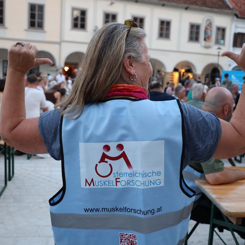 Eine Frau mit einer blauen Weste mit dem Text 'sterreische MUSKELFORSCHUNG' und einem Logo steht in einem Außenbereich, möglicherweise bei einer Veranstaltung. Sie zeigt mit den Fingern nach oben. Hinter ihr sitzen Menschen auf Stühlen und Tischen. In der Ferne befindet sich ein Gebäude.