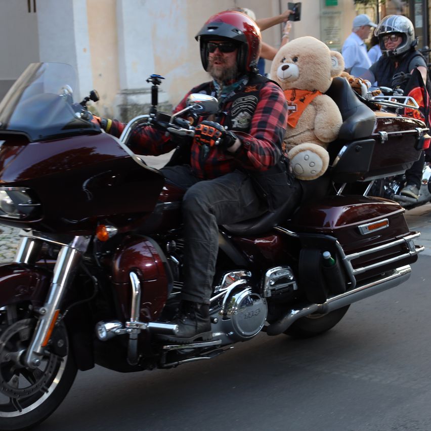 Ein Mann fährt auf einem Motorrad mit einem großen Teddybären auf seinem Schoß. Er trägt einen roten Helm und Sonnenbrille. Hinter ihm folgt ein weiterer Motorradfahrer.