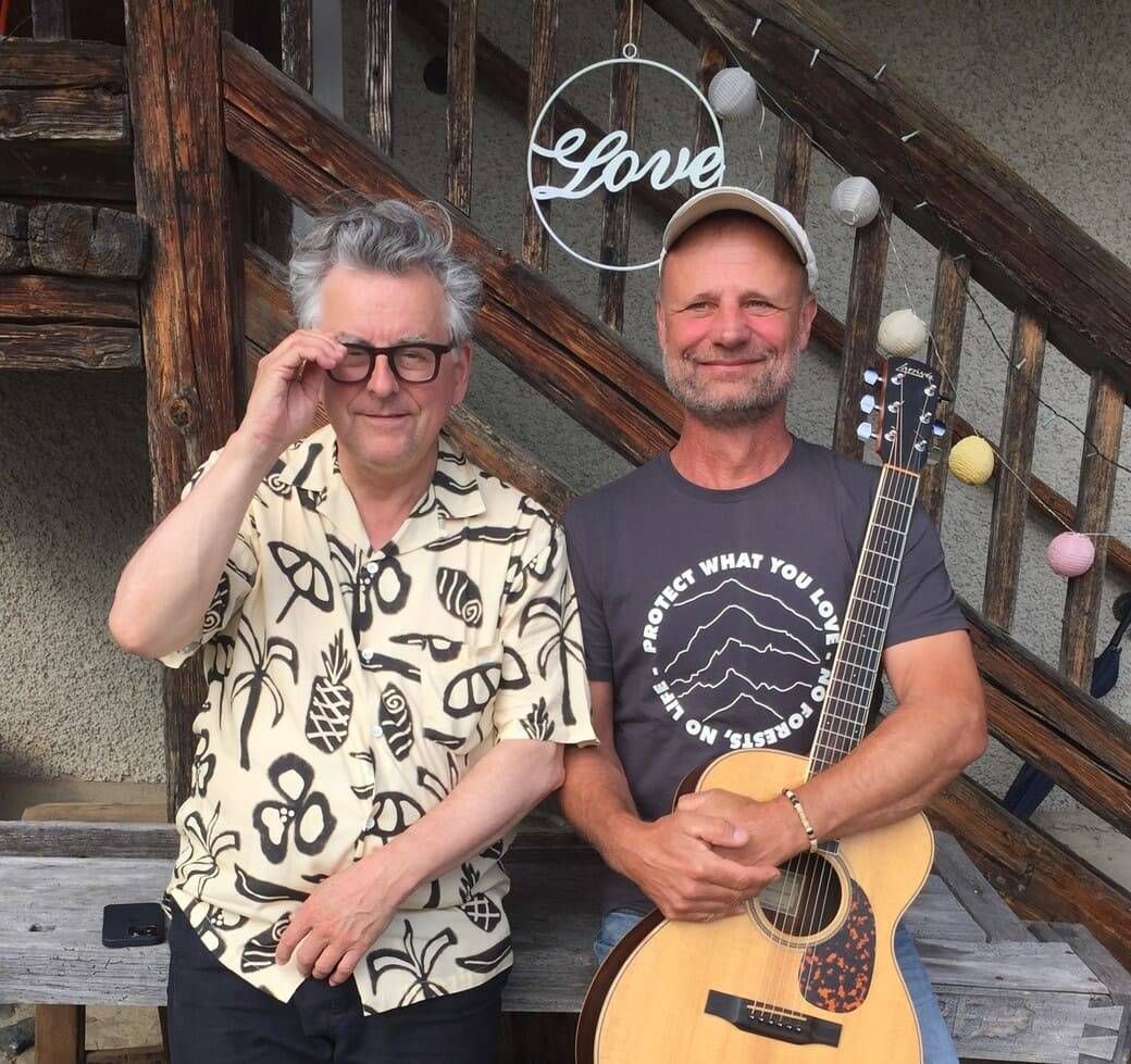 Zwei Männer stehen auf einer Holztreppe, einer hält eine Gitarre und lächelt, mit einem dekorativen Schild über ihnen.