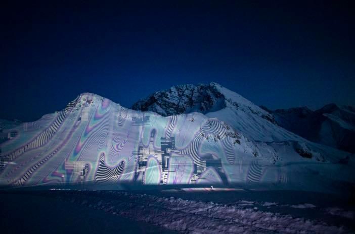 Ein nächtlicher Blick auf einen Berg mit einer farbenfrohen, abstrakten Lichtprojektion auf seiner Oberfläche. Die Projektion wird von einer Lichtquelle beleuchtet und wirft einen Schatten auf den verschneiten Boden.