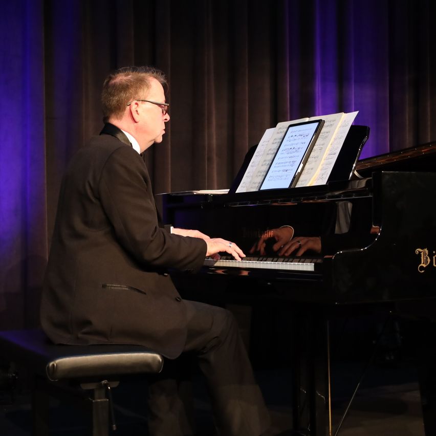 Ein Mann sitzt auf einer Bank und spielt auf der Bühne Klavier mit geöffneten Notenblättern vor sich.