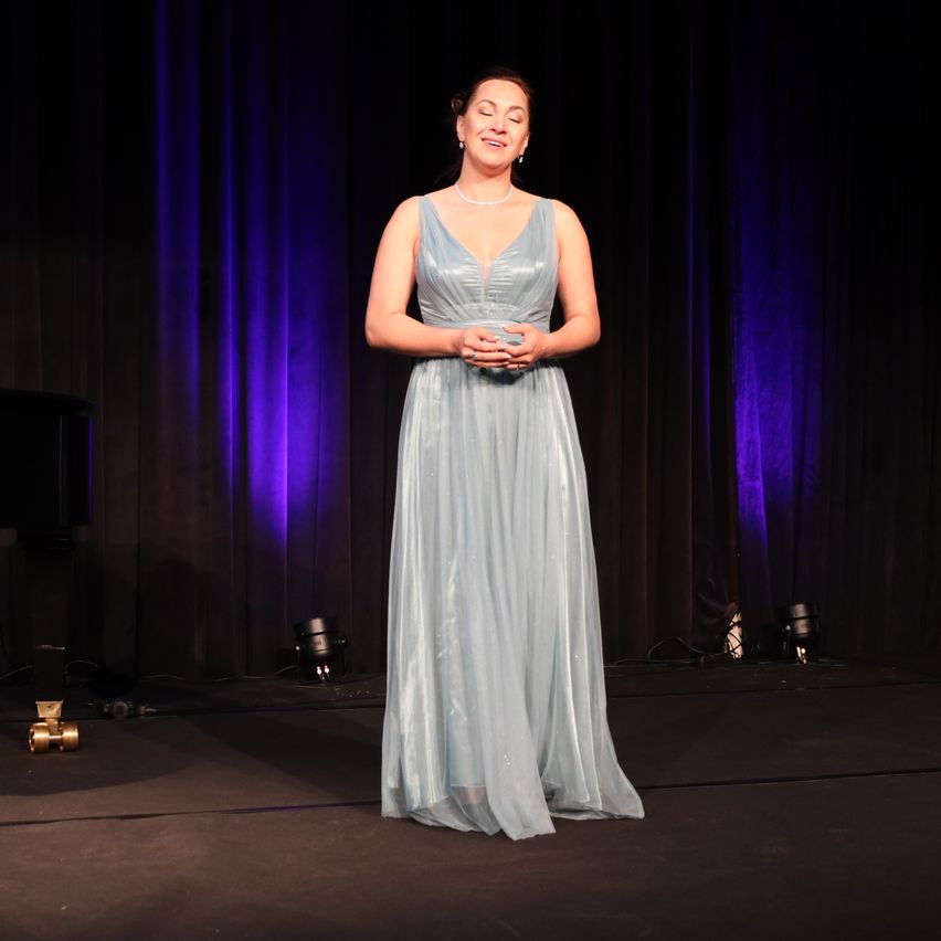 Eine Frau in einem hellblauen Kleid steht auf der Bühne mit einem Klavier im Hintergrund, beleuchtet von lila Lichtern.