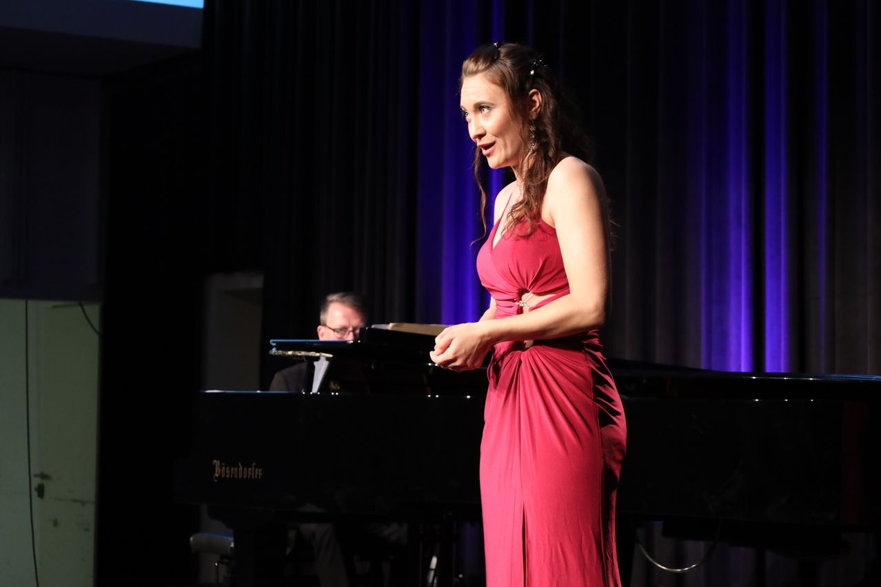 Eine Frau in einem roten Kleid steht auf der Bühne neben einem Klavier, ein Mann spielt im Hintergrund. Der Hintergrund ist dunkel mit blauen Vorhängen.