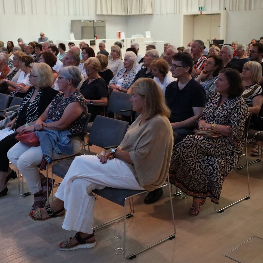 Ein Raum voller Menschen, die auf Stühlen sitzen. Die meisten von ihnen sind ältere Frauen und Männer, die einer Präsentation zuhören. Einige tragen Brillen und Armbänder. Der Raum hat weiße Wände und Holzböden.
