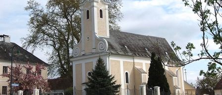 Eine Kirche mit hohem Turm steht neben einer Straße, davor Bäume und ein Schild mit der Aufschrift 'Weinarchiv'. Im Vordergrund befinden sich eine Straßenlaterne und ein Zaun, links steht ein Gebäude.