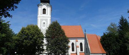 Eine weiße Kirche mit rotem Dach und einem hohen Turm steht in einem Friedhof, umgeben von Bäumen und Gras.
