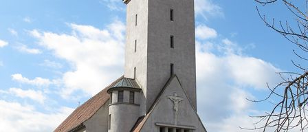 Eine Kirche mit einem Turm, einem braunen Dach und einer Statue von Jesus an der Fassade. Zwei Statuen flankieren den Eingang.