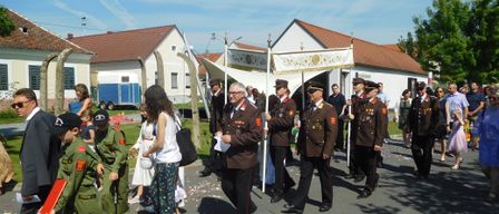Ein Militäraufmarsch vor einem Haus, Menschen in Uniformen und Kinder in grünen Outfits, einer hält einen Fahnenmast, ein weißes Zelt im Hintergrund und Menschen, die auf einer Straße gehen.